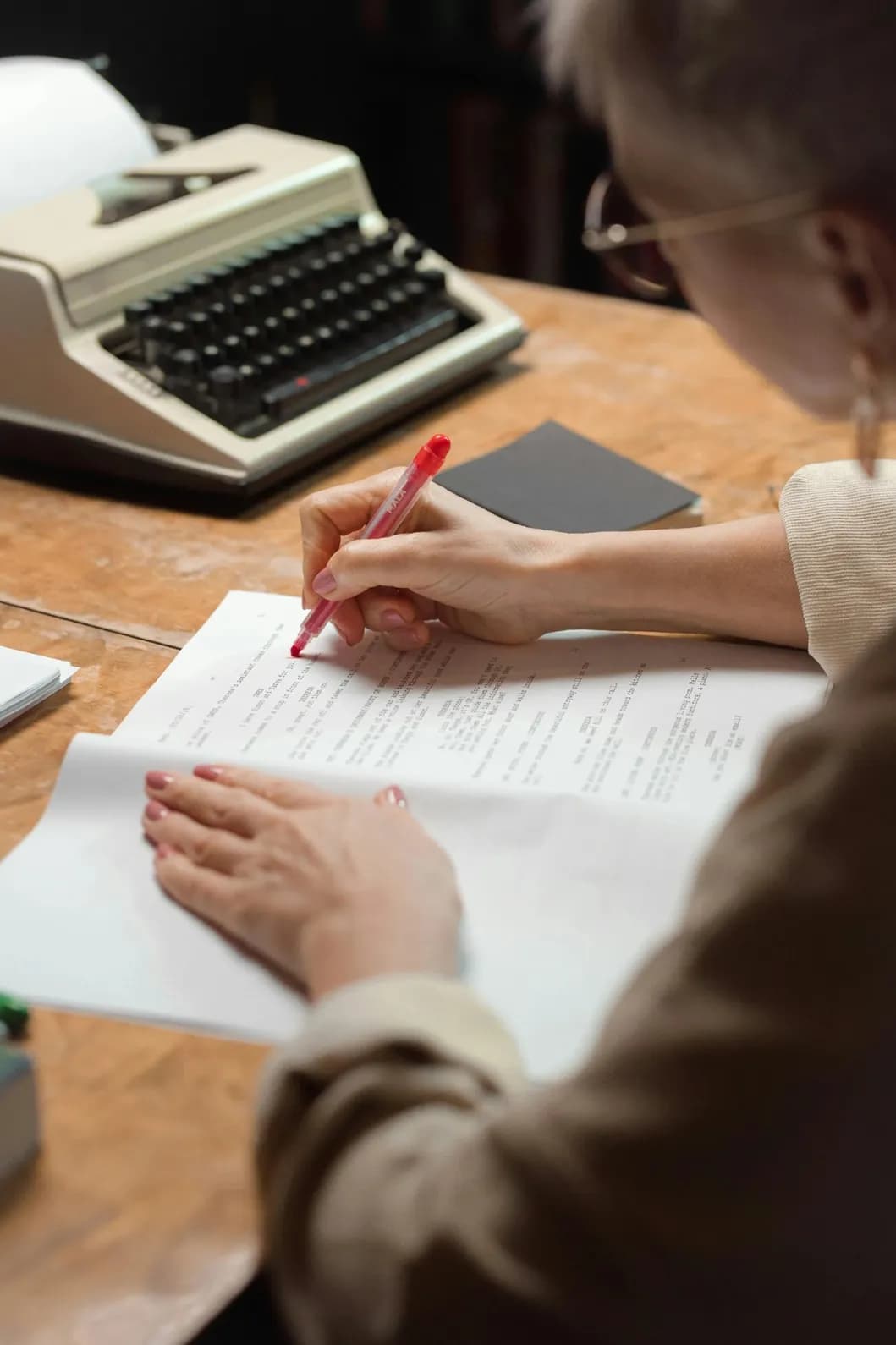 Person editing a document with a red pen while a typewriter is placed on the desk, focusing on text revisions.